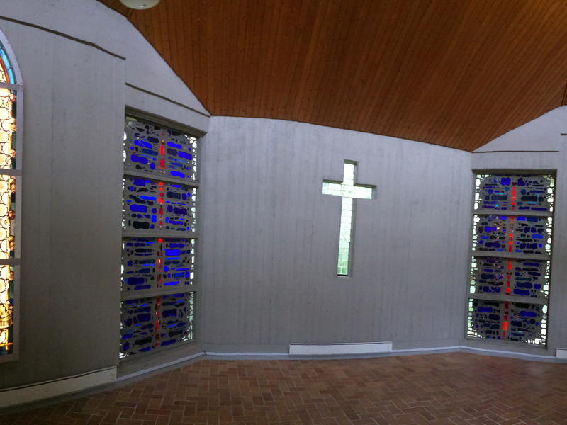 Bild des Friedhofgebäudes von innen, zu sehen in in der Mitte der Wand ein Kreuz auf beiden Seiten nebenan sind bunte Glasbausteine und auf beiden Seiten daneben sind beleuchtete Figuren