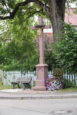 Bild eines großen Jesus-Kreuzes und einer Sitzbank vor einem Zaun und einem Baum