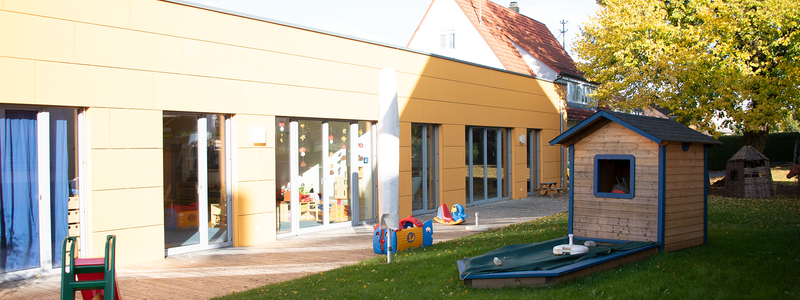 Bild des hinteren Bereichs des Kindergartens mit einem Teil des Gartens, im Garten befindet sich ein kleines Gartenhaus und eine Rutsche