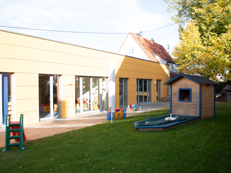Bild des hinteren Bereichs des Kindergartens mit einem Teil des Gartens, im Garten befindet sich ein kleines Gartenhaus und eine Rutsche
