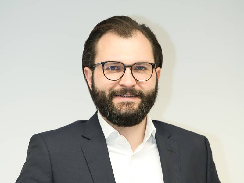 Ein Mann mit Bart und Brille trägt einen dunklen Anzug und ein weißes Hemd vor einem neutralen Hintergrund.