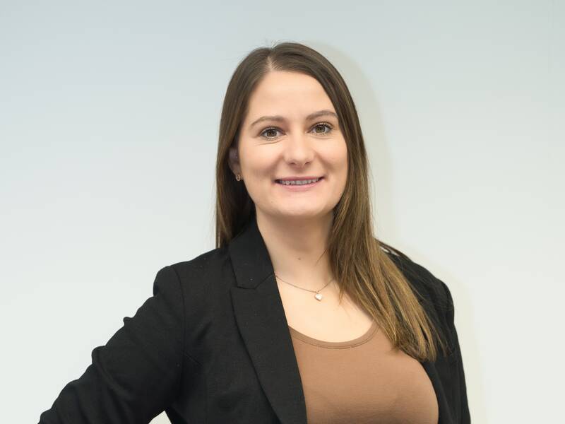Eine lächelnde Frau mit braunen Haaren trägt einen schwarzen Blazer und ein braunes Oberteil vor einem neutralen Hintergrund.