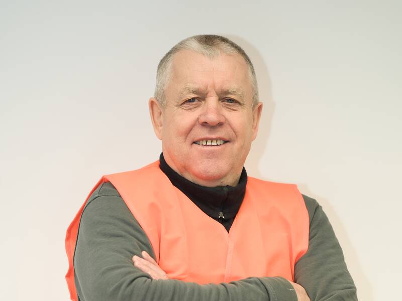 Ein Mann mit kurzen grauen Haaren trägt eine orangefarbene Warnweste und hat die Arme verschränkt vor einem hellen Hintergrund.