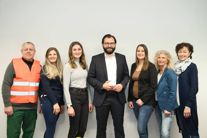 Sieben Personen stehen lächelnd nebeneinander vor einem neutralen Hintergrund, wobei eine Person eine orangefarbene Warnweste trägt.