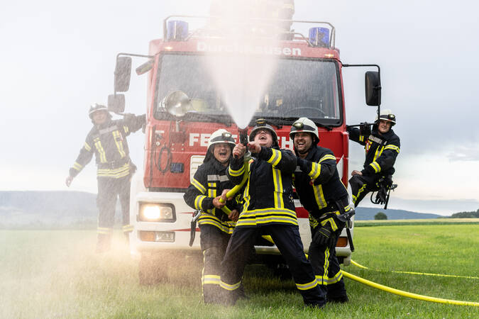 Fünf Personen der Feuerwehr, in der Mitte steht das Löschfahrzeug, zwei Feuerwehrmänner halten sich rechts und links am Fahrzeug und drei Feuerwehrmänner stehen vor dem Fahrzeug mit dem Feuerwehrschlauch in der Hand