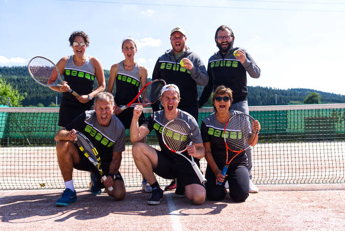 Bild von sieben Personen des Sportvereins der Abteilung Tennis, welche auf dem Tennisplatz vor dem Netz stehe und teilweise den Tennisschläger und eine starke Faust zeigen