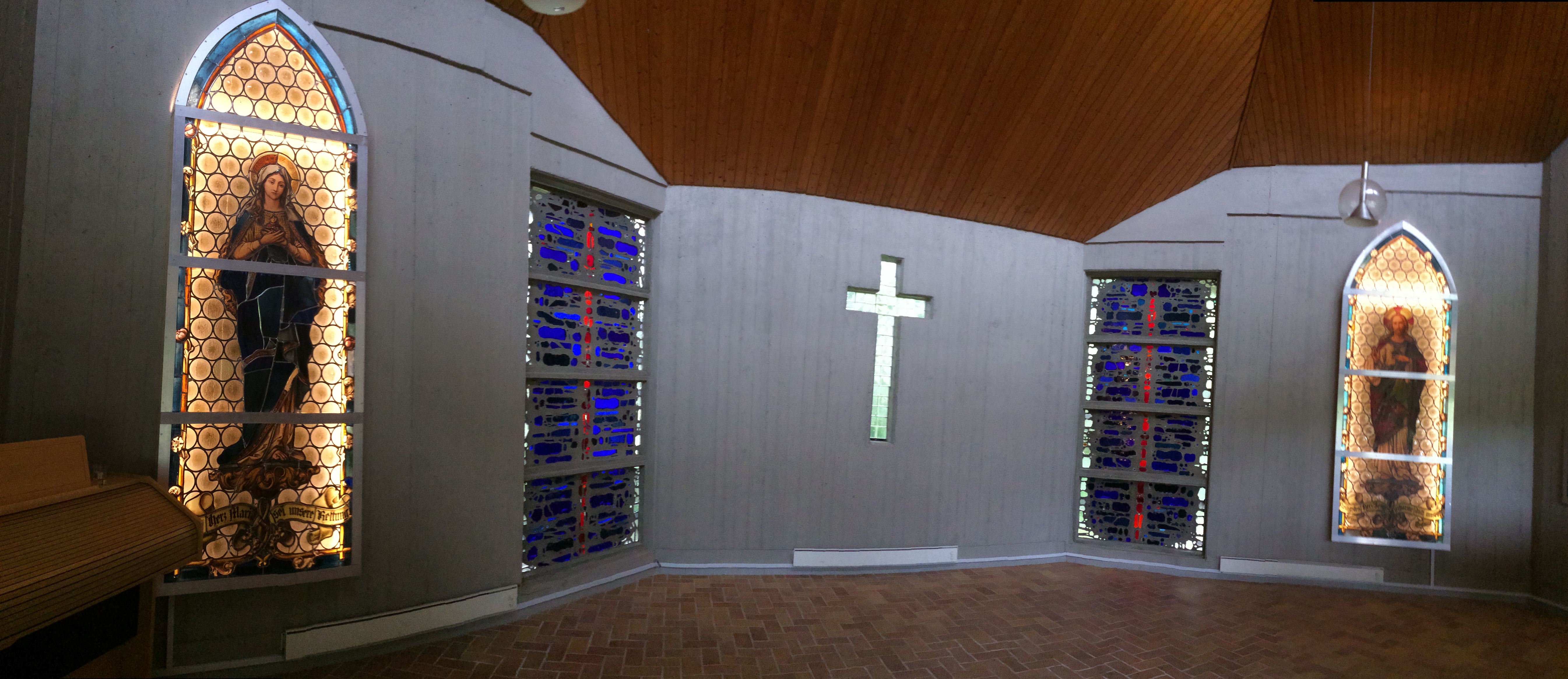 Bild des Friedhofgebäudes von innen, zu sehen in in der Mitte der Wand ein Kreuz auf beiden Seiten nebenan sind bunte Glasbausteine und auf beiden Seiten daneben sind beleuchtete Figuren