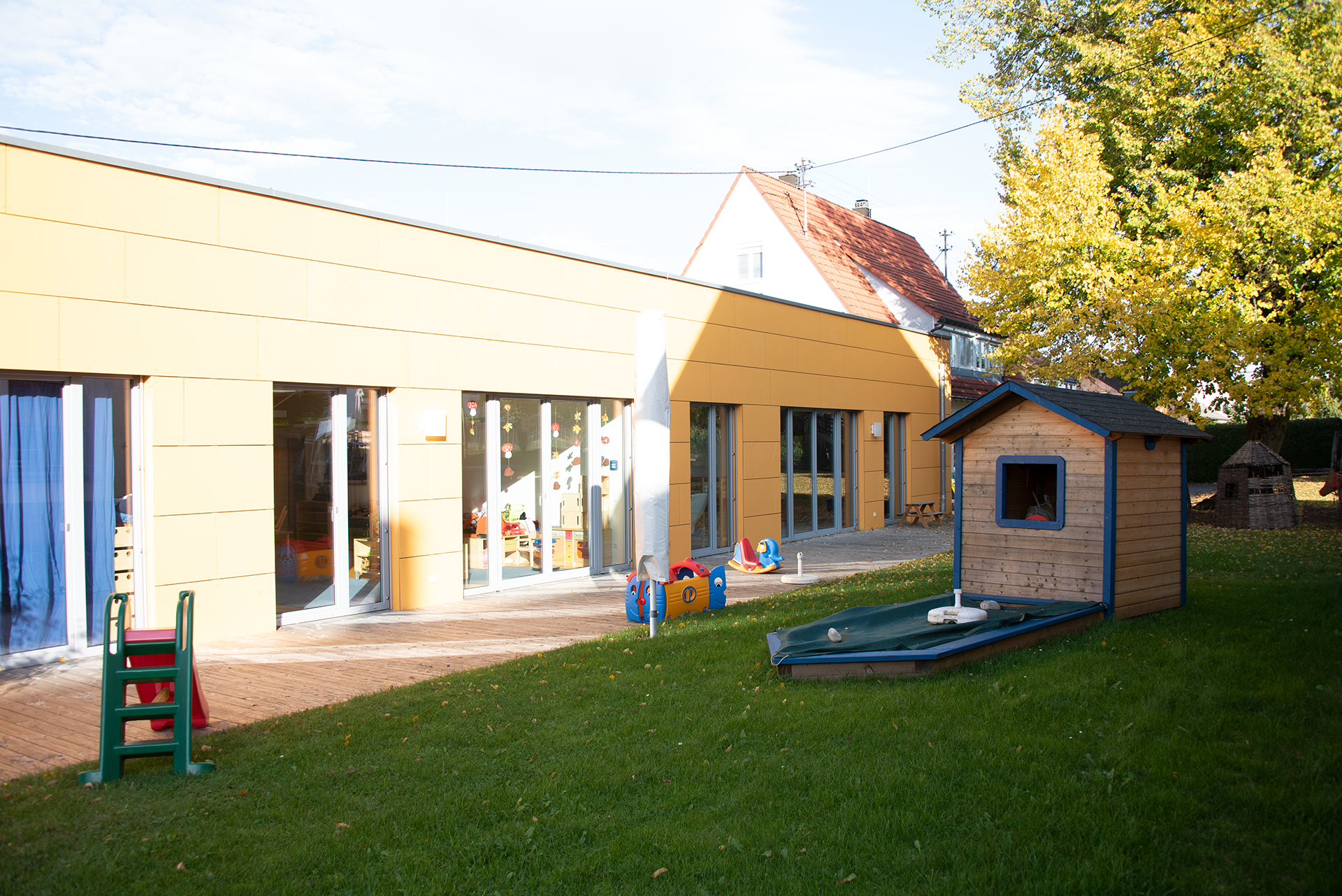 Bild des hinteren Bereichs des Kindergartens mit einem Teil des Gartens, im Garten befindet sich ein kleines Gartenhaus und eine Rutsche